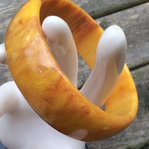 May include: A marbled, amber-coloured bangle bracelet. The thick, rounded bracelet has a glossy finish and a warm, golden hue with streaks of darker brown. The bracelet is displayed on a white, stylised hand.