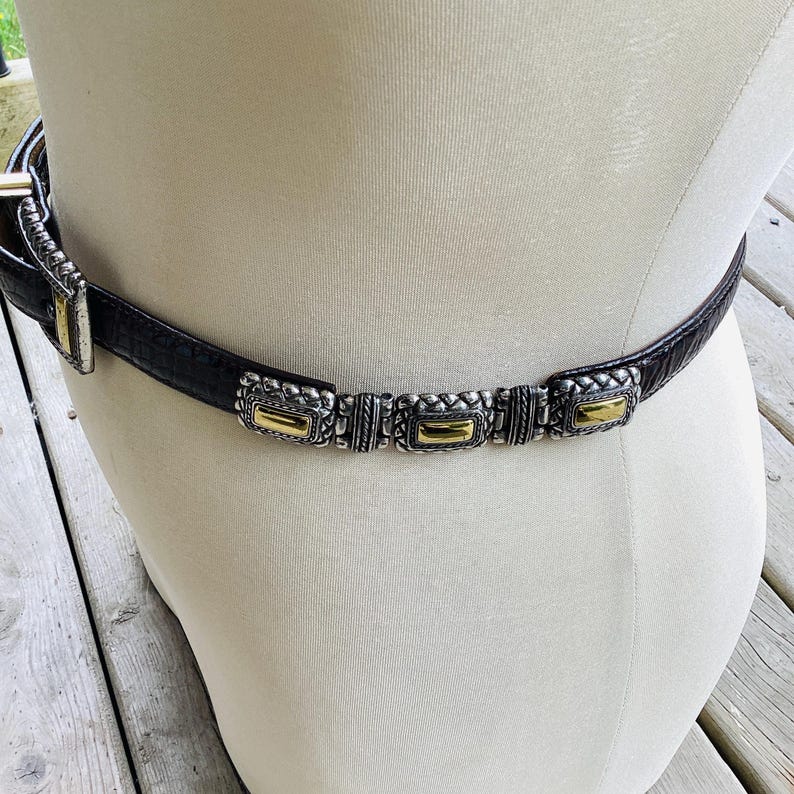 May include: Black belt with silver and gold rectangular details. The belt is made of a textured material and has a silver buckle. The decorative details are evenly spaced along the belt's length, adding a touch of elegance.