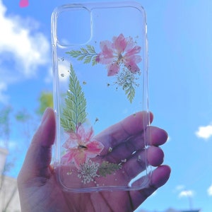 May include: A clear phone case with pressed pink and white flowers and greenery inside. The case is held in a hand against a blue sky background.
