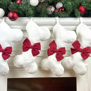 Op de afbeelding: Vier witte gewatteerde kerstsokken met rode strikken hangen aan een witte schoorsteenmantel. De sokken zijn botvormig en hebben een ruitpatroon. De schoorsteenmantel is versierd met groen, rode en witte ornamenten.