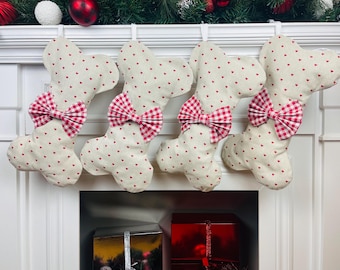 May include: Four dog bone-shaped Christmas stockings. The stockings are cream-coloured with small red hearts and a red and white gingham bow. They are hanging above a fireplace, decorated with Christmas ornaments.