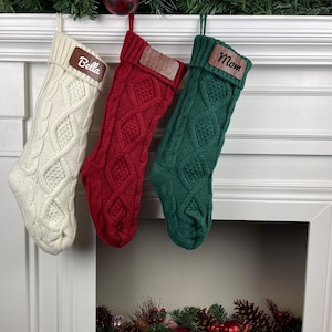 May include: Three knitted Christmas stockings with personalised wooden name tags. The stockings are white, red, and green with a cable knit pattern. The names on the tags are "Bella", "Mom", and "Mom".