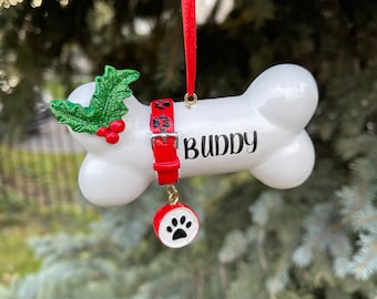 May include: A white bone-shaped Christmas ornament with the name "Buddy" in black. It features a red collar with paw prints and a small charm with a paw print. Green holly and red berries are on one end. A red ribbon is attached for hanging.