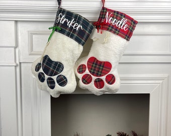 May include: Two white Christmas stockings with red and green plaid trim and paw print designs. The stockings are personalized with the names "Striker" and "Noodle".