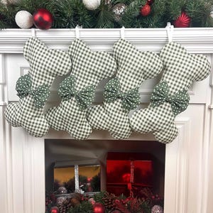 Peut inclure: Quatre bas de Noël avec un motif vichy vert et blanc et un nœud vert à pois. Les bas sont suspendus au-dessus d'une cheminée décorée d'ornements et de verdure de Noël.
