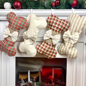 May include: Four dog bone-shaped Christmas stockings. Two are plaid with red, green, and white, one is solid cream, and one is striped. Each has a burlap-style band and a cream bow. The stockings hang above a fireplace.