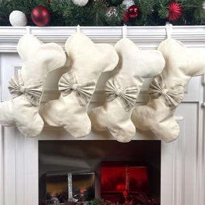 May include: Four cream-coloured Christmas stockings shaped like dog bones, each with a plaid bow. They hang above a fireplace decorated with festive greenery and baubles, creating a cosy holiday scene.