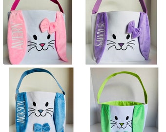 Puede incluir: Cuatro cestas de Pascua de conejito personalizadas con orejas y asas de diferentes colores. Las cestas son blancas con una cara de conejito y un lazo. Los nombres Avery, Sawyer, Jackson y un cuarto nombre están bordados en las orejas.