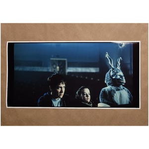 Puede incluir: Una impresi&oacute;n enmarcada de una escena de pel&iacute;cula. Tres personas est&aacute;n sentadas, una de ellas con un gran disfraz de conejo con una expresi&oacute;n amenazante. El fondo es oscuro, con un tono azul, lo que sugiere una escena nocturna.