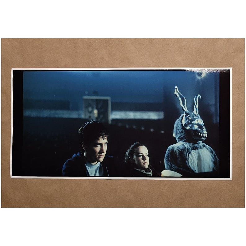 Puede incluir: Una impresi&oacute;n enmarcada de una escena de pel&iacute;cula. Tres personas est&aacute;n sentadas, una de ellas con un gran disfraz de conejo con una expresi&oacute;n amenazante. El fondo es oscuro, con un tono azul, lo que sugiere una escena nocturna.