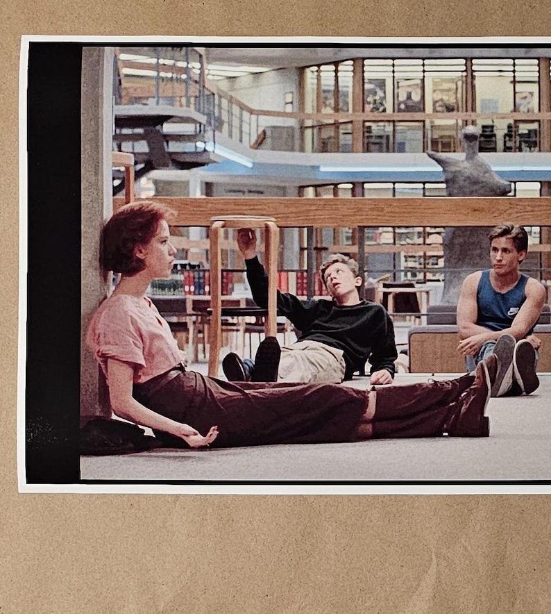May include: A framed print featuring a scene from a movie. Three people are in a library setting. One person is sitting on the floor, wearing a pink top and brown skirt. Two other people are sitting nearby. The background shows bookshelves and architectural details.