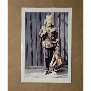 May include: A full-length photograph of a person holding a long, cylindrical object and a large canvas bag. The person is wearing a dark jacket, dark pants, and light-colored shoes. The background is a dark, vertical-paneled wall.