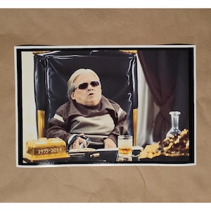 May include: A framed print featuring a person wearing sunglasses and a brown sweater, seated in a black leather chair. A glass of amber liquid, a decanter, and a box with the text "1975-2014" are on the table.