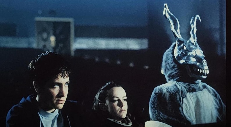 Puede incluir: Una escena de una pel&iacute;cula con tres personas. Una persona lleva un amenazante disfraz de conejo con una cara esquel&eacute;tica. Las otras dos personas miran fuera de la pantalla con expresiones serias. El fondo es oscuro.