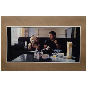 May include: A framed photograph featuring a man and a teddy bear seated on a dark sofa. A table in front holds snacks, a bowl, a glass of juice, and a cereal box. The teddy bear is light brown.