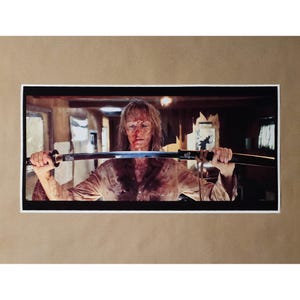 May include: A framed photograph featuring a person holding a katana sword. The person is covered in blood and wearing a button-down shirt. The background shows a dimly lit interior with windows and wooden elements.