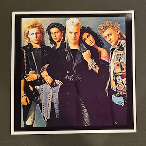 May include: A square print featuring a group of six people. The image has a dark, stylized aesthetic. The subjects are dressed in dark clothing, with some wearing leather jackets. The image has a black border and a white background.
