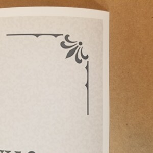 May include: A corner detail of a printed page featuring a decorative gray floral design. The design is composed of stylized leaves and a central dot, with a border of short vertical lines. The paper has a textured, off-white appearance.
