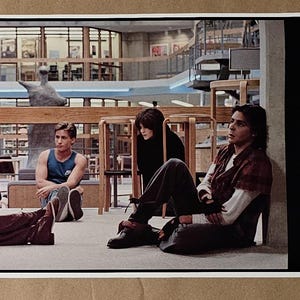May include: A still from a movie featuring three people in a library setting. One person wears a blue tank top, another a black top, and the third a red plaid shirt. The scene is set in a library with wooden furniture.