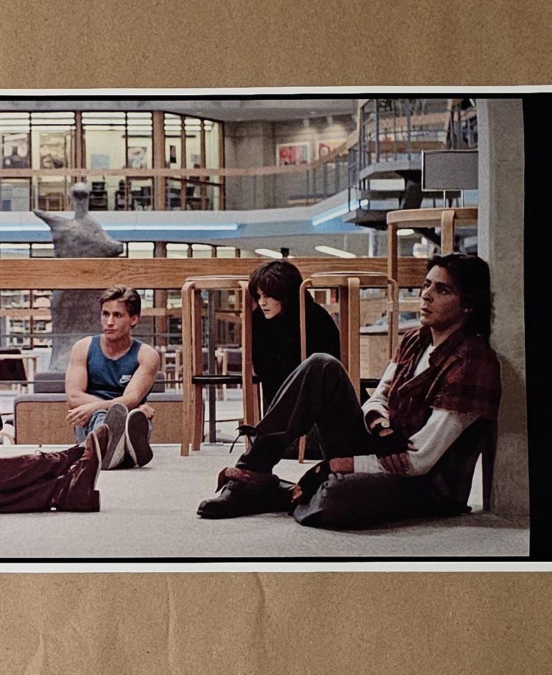 May include: A still from a movie featuring three people in a library setting. One person wears a blue tank top, another a black top, and the third a red plaid shirt. The scene is set in a library with wooden furniture.