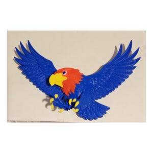 May include: A three-dimensional wall-mounted eagle figure featuring a vibrant blue body and wings, a bright orange head, and a yellow beak and talons. The eagle is depicted in a dynamic flying pose with wings fully extended and claws forward.