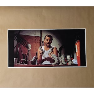 May include: A framed print featuring a man in a white tank top holding several small, gray dinosaur sculptures. The scene is set in a dimly lit workshop with various tools and a fire extinguisher visible.