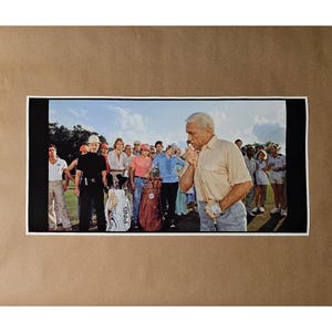 May include: A framed print featuring a group of people on a golf course. A man in a light yellow shirt and gray pants is in the foreground, holding a golf club. Other people are in the background, some wearing golf attire. Golf bags are visible.