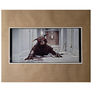 May include: A framed print featuring a menacing creature with red eyes and sharp teeth, crawling in a hallway. The creature has horns and claws. The hallway has white walls and a patterned wallpaper in the background. The print is set against a brown background.