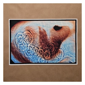 May include: Abstract art print featuring concentric circles and rippling water effects in shades of blue and brown. The artwork is framed with a white mat and a black border, set against a brown background.