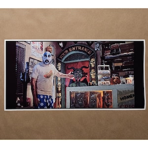 May include: A framed print featuring a man in clown makeup standing in front of a tour entrance. The entrance is decorated with skulls and a devil figure. The man is wearing a t-shirt with a hot dog graphic.