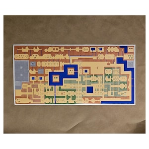 May include: A rectangular map print featuring a pixelated design with tan, blue, green, and brown elements. The map depicts a detailed layout of a fictional landscape, with various structures and pathways. The print is on a white background.