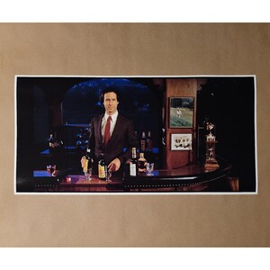May include: A framed print featuring a man in a suit standing behind a bar with bottles and glasses. The bar has a dark wood finish. There are framed pictures on the wall behind the bar. The print is on a brown background.