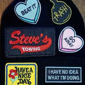 May include: A black beanie with various colorful embroidered patches. The patches include a red heart with the text "Beat It", a yellow trash bag with the text "Trash", a red and white heart with the text "Go Away", a black patch with the text "Steve's Towing", a yellow patch with a flower and the text "Have a Nice Day", and a gray patch with the text "I Have No Idea What I'm Doing".