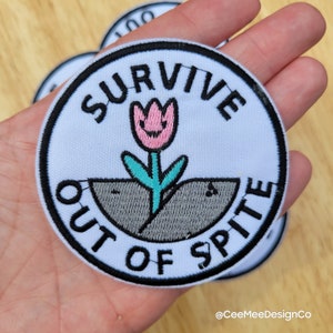 May include: White embroidered patch with black stitching. The patch is round and features a pink flower with a smiling face growing out of gray ground. The text "SURVIVE OUT OF SPITE" is written in a black circular border around the flower.