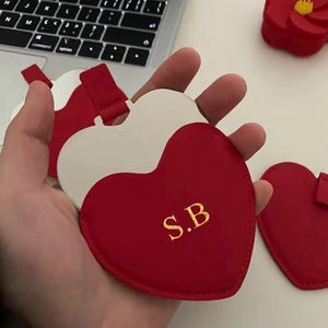 May include: Heart-shaped red and white leather luggage tags with gold initials "S.B". The tags have a red strap and are likely used for personalising luggage or bags. The design is simple and elegant.