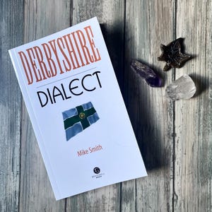 May include: A book titled "Derbyshire Dialect" by Mike Smith, with a flag design on the cover. The book is placed on a wooden surface, alongside several crystals. The title is in a bold, orange font.