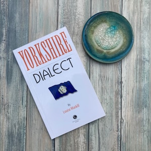 May include: A white book titled "Yorkshire Dialect" by Louise Maskill. The book features a blue and white flag with a white rose in the centre. The book is on a wooden surface with a blue and green ceramic dish.