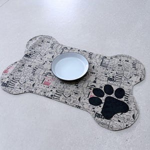 Peut inclure: Un tapis de nourriture pour animaux de compagnie en forme d'os avec un bol en céramique blanc. Le tapis présente un motif répété de chats de dessins animés et une empreinte de patte noire. Le bol est centré sur le tapis.