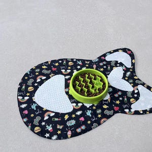 Peut inclure: Tapis de nourriture pour animaux de compagnie noir en forme de poisson, orné d'icônes de dessins animés colorés. Un bol vert pour alimentation lente rempli de croquettes marron est posé sur le tapis.