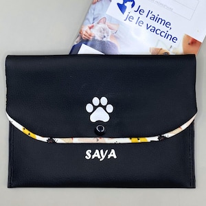 Peut inclure: Pochette noire pour documents d'animaux avec une empreinte de patte blanche et le nom "SAYA" imprimé en blanc. La pochette a une bordure à motifs et une fermeture à bouton. Un document avec du texte français est partiellement visible.