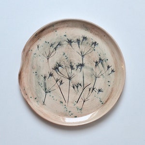 Op de afbeelding: Een keramisch bord met een lichtbruine glazuur en een ontwerp van zwarte wilde bloemen. Het bord heeft een licht ongelijke rand.