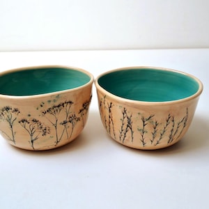 Pode incluir: Dois recipientes de cerâmica com um exterior bege claro liso, apresentando ilustrações botânicas escuras e detalhadas de flores silvestres. O interior de cada recipiente tem um acabamento em esmalte turquesa sólido e vibrante.
