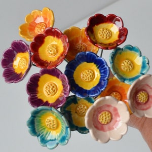 May include: A collection of ceramic flowers on thin metal stems, each featuring a glossy finish and a textured yellow center. The petals display a variety of vibrant colors including red, orange, purple, blue, teal, and soft pink, arranged in a cluster.