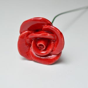 May include: A handcrafted, glossy red ceramic rose with detailed petals. The flower is on a green wire stem, set against a white background. The rose is a decorative item.