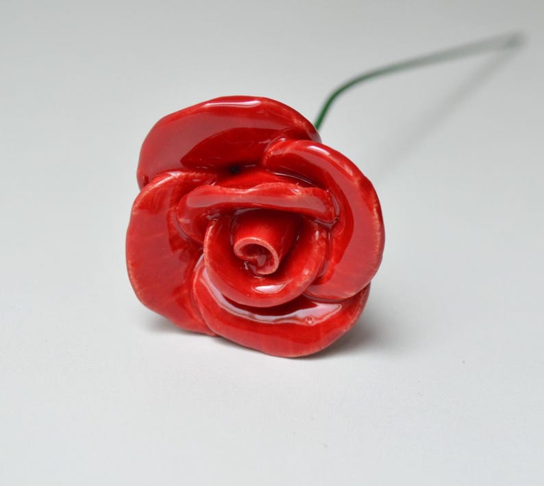 May include: A handcrafted, glossy red ceramic rose with detailed petals. The flower is on a green wire stem, set against a white background. The rose is a decorative item.