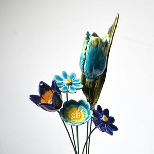Può includere: Un bouquet di fiori in ceramica in tonalità di blu, giallo e verde. I fiori sono disposti su steli di filo metallico e comprendono un tulipano, una margherita e altri due tipi di fiori.