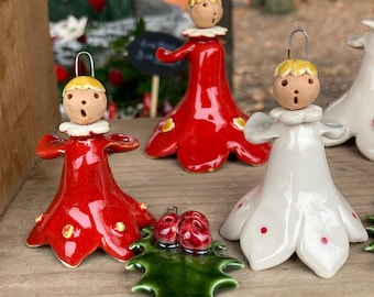 Peut inclure: Trois décorations de Noël en céramique en forme d'anges. Deux sont rouges avec des accents jaunes et une est blanche avec des accents rouges. Les décorations sont sur une surface en bois avec une feuille de houx verte.