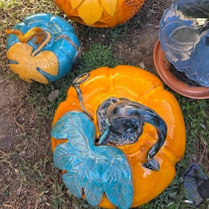 Könnte beinhalten: Drei Keramikkürbisse mit glänzender Oberfläche. Einer ist leuchtend orange mit dunklem Stiel und türkisfarbenem Blatt, ein anderer ist türkis mit gelbem Blatt und Stiel, und der dritte ist orange mit einem gelben Blatt. Jeder hat einen Stiel.