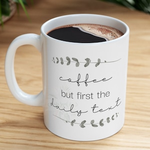 May include: White ceramic coffee mug with black text that reads "coffee but first the daily text" and a floral design in grey.