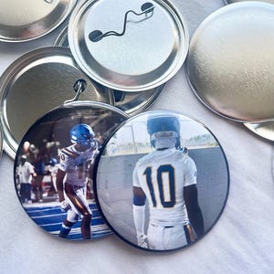 May include: A collection of round pinback buttons featuring images of football players in action. The buttons display players in white and blue uniforms, with the number 10 prominently visible. The buttons are silver-colored on the back.
